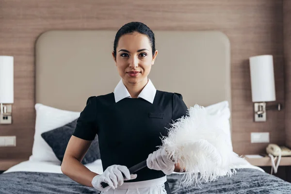 Front view of smiling maid in white gloves holding duster near bed and looking at camera — Stock Photo