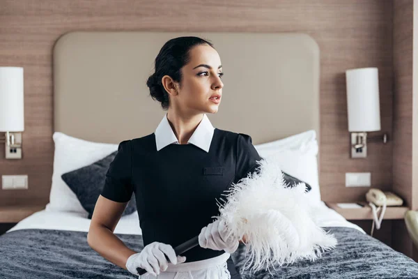 Nachdenkliches brünettes Dienstmädchen in Schürze und Handschuhen, Staubwedel haltend und im Hotelzimmer wegschauend — Stockfoto