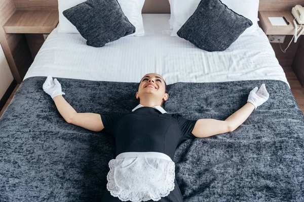 Empregada sorridente em avental branco e luvas deitadas na cama no quarto do hotel — Fotografia de Stock