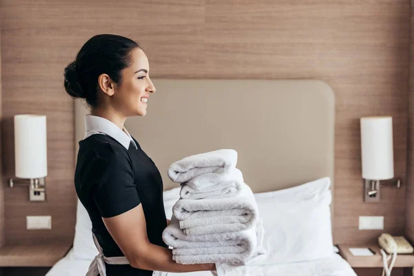 Vista laterale di cameriera sorridente che tiene mucchio di asciugamani piegati vicino al letto in camera d'albergo — Foto stock