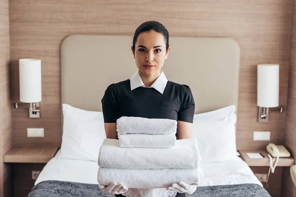 Vue de face de la bonne tenant pile de serviettes pliées près du lit et regardant la caméra dans la chambre d'hôtel — Photo de stock