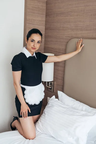 Empregada sorridente em avental branco em pé perto da cama e olhando para a câmera no quarto do hotel — Fotografia de Stock