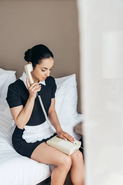 Bonita sirvienta en delantal blanco sentada en la cama y hablando por teléfono en la habitación de hotel - foto de stock