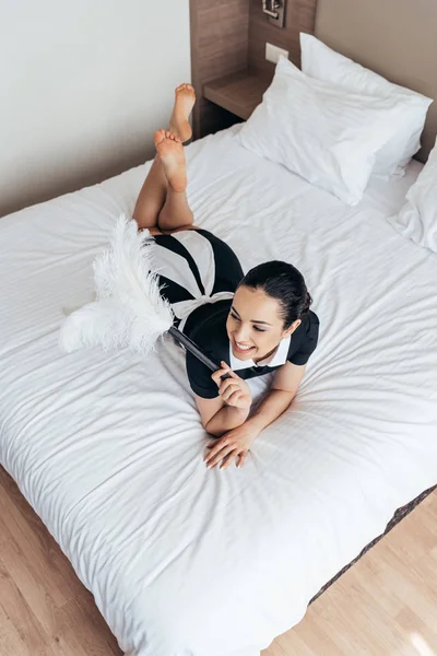 Vue aérienne de souriante femme de ménage pieds nus couchée sur le lit et tenant plumeau dans la chambre d'hôtel — Photo de stock