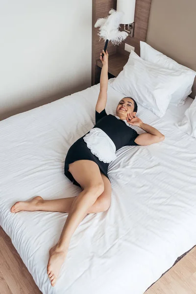 Улыбающаяся босиком горничная лежит на кровати и держит пыльник в гостиничном номере — стоковое фото