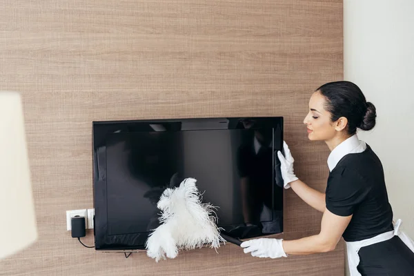 Pretty maid in white gloves cleaning tv with duster — Stock Photo