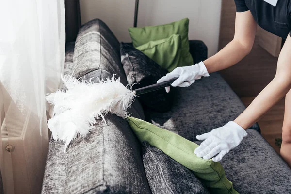 Vista parcial de la mucama en guantes blancos limpieza sofá con plumero en habitación de hotel - foto de stock