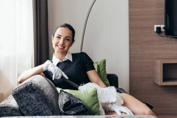 Sorrindo empregada doméstica em luvas brancas sentado no sofá e segurando espanador no quarto do hotel — Fotografia de Stock