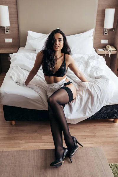 Sexy criada en negro medias y blanco delantal sentado en la cama y mirando a la cámara - foto de stock