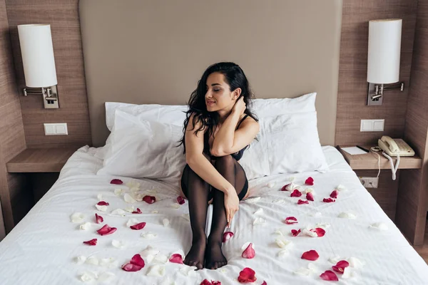 Sexy fille brune en bas noirs assis sur le lit avec des pétales de rose dans la chambre — Photo de stock