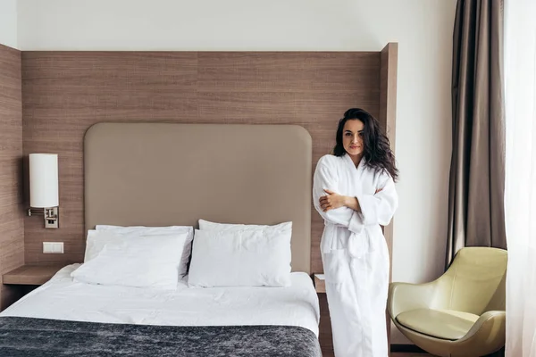 Attractive brunette young woman in white bathrobe standing near bed in cozy bedroom — Stock Photo