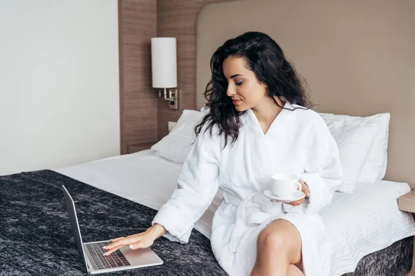 Affascinante bruna giovane donna in bagno bianco utilizzando il computer portatile e tenendo in mano una tazza di caffè in camera da letto — Foto stock