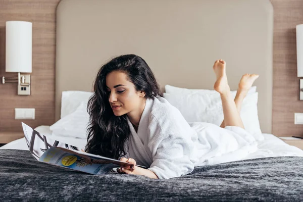 Bonita morena descalza joven en albornoz blanco acostada en la cama y leyendo la revista - foto de stock