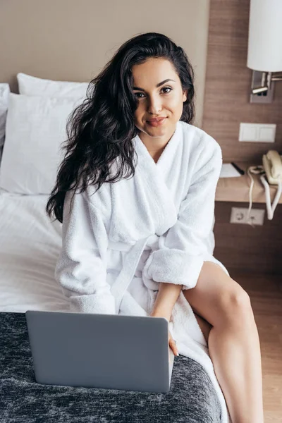 Encantadora morena joven en el baño blanco utilizando el ordenador portátil en el dormitorio - foto de stock