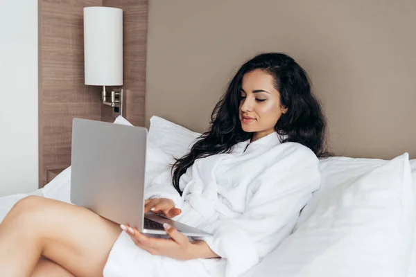 Affascinante bruna giovane donna in bagno bianco utilizzando il computer portatile in camera da letto — Foto stock