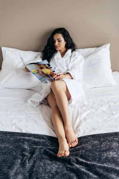Jolie brune jeune femme en peignoir blanc couché sur le lit et lecture magazine — Photo de stock