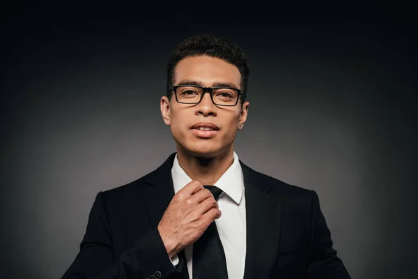 Handsome african american businessman in glasses and suit touching tie on dark background — Stock Photo