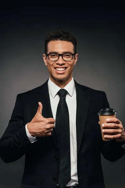 Красивый улыбающийся африканский бизнесмен в очках и костюме держит кофе, чтобы пойти и показать большой палец на темном фоне — стоковое фото