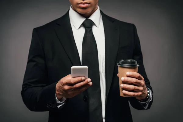 Vue recadrée de l'homme d'affaires afro-américain tenant du café à emporter et smartphone sur fond sombre — Photo de stock