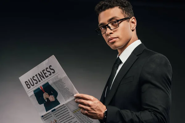 Side view of african american businessman in glasses and suit reading business newspaper on dark background — Stock Photo