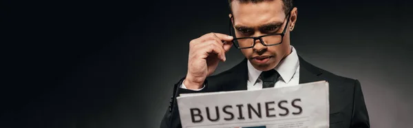 Guapo hombre de negocios afroamericano en gafas y traje leyendo periódico de negocios sobre fondo oscuro, plano panorámico - foto de stock