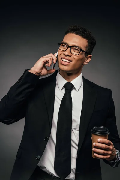 Handsome african american businessman with coffee to go talking on smartphone on dark background — Stock Photo