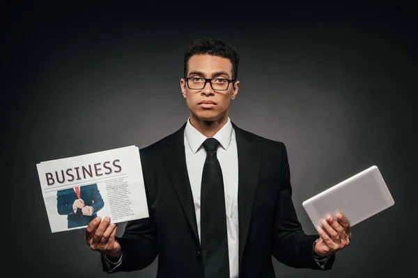 African american businessman in glasses and suit holding business newspaper and digital tablet on dark background — Stock Photo