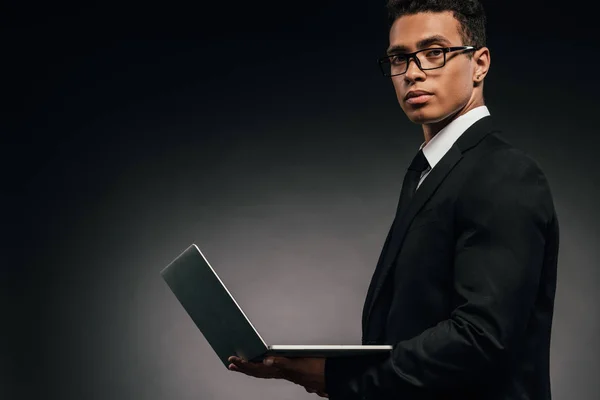 Vue latérale de l'homme d'affaires afro-américain tenant un ordinateur portable sur fond sombre — Photo de stock