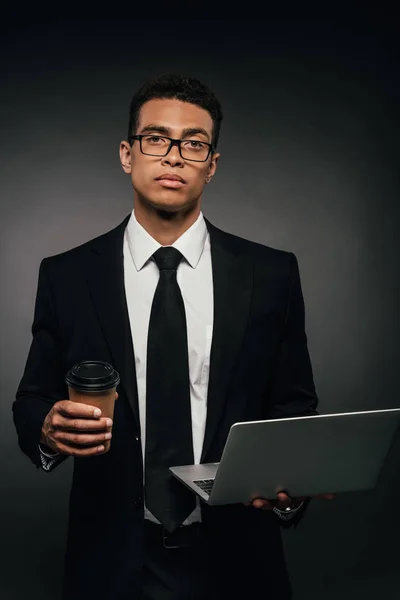 African american businessman holding laptop and paper cup on dark background — Stock Photo