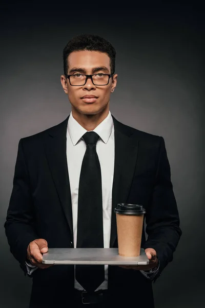 Homme d'affaires afro-américain dans des lunettes tenant ordinateur portable et tasse en papier sur fond sombre — Photo de stock