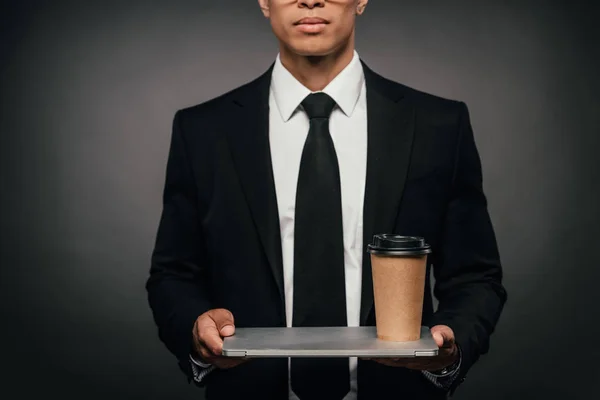Cropped view of african american businessman holding laptop and paper cup on dark background — Stock Photo