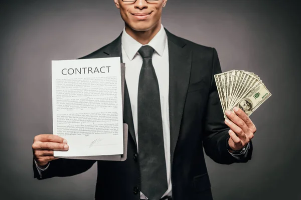 Обрізаний вид щасливого афроамериканського бізнесмена, що показує контракт і тримає готівку на темному тлі — стокове фото