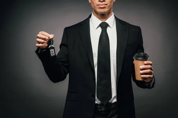 Vista parcial del hombre de negocios afroamericano sosteniendo las llaves del coche y la taza de papel sobre fondo oscuro - foto de stock