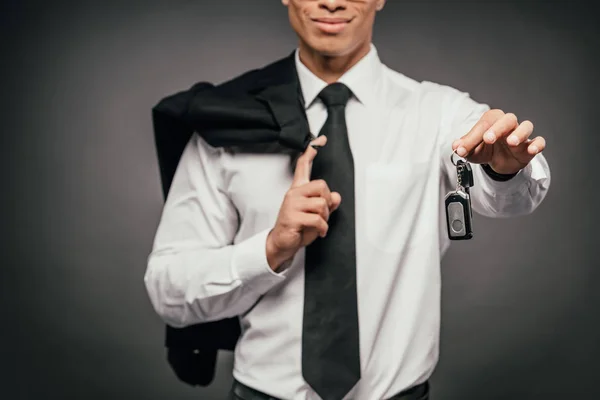 Vista ritagliata di sorridente uomo d'affari africano americano in possesso di blazer, chiavi di auto su sfondo scuro — Foto stock