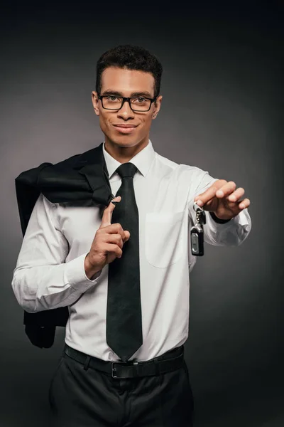 Sonriente hombre de negocios afroamericano sosteniendo la chaqueta y las llaves del coche sobre fondo oscuro - foto de stock