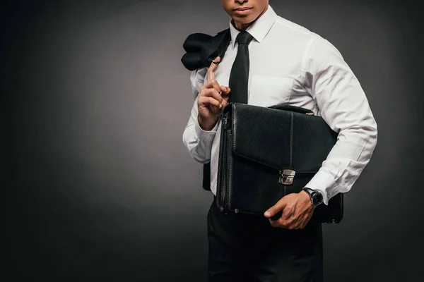 Cropped view of african american businessman holding blazer and leather briefcase on dark background — Stock Photo