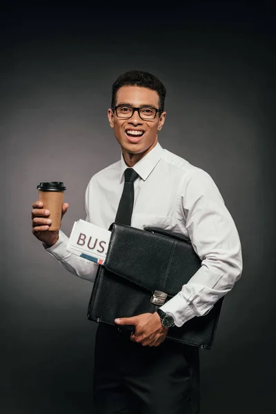 Happy african american businessman holding newspaper, coffee to go and leather briefcase on dark background — Stock Photo