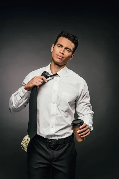 Hombre de negocios afroamericano con dinero en efectivo en el bolsillo sosteniendo café para ir y desatar la corbata sobre fondo oscuro - foto de stock