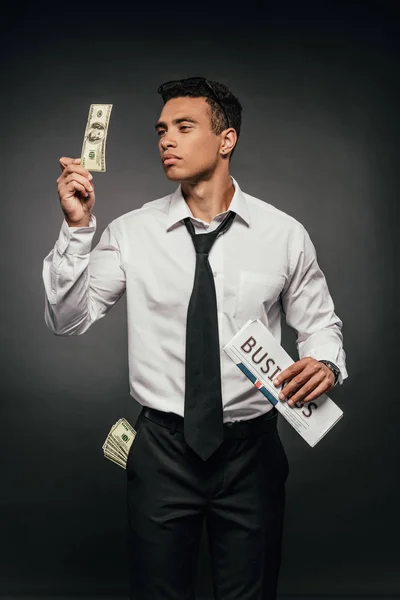 Wealthy african american businessman holding business newspaper and looking at banknote on dark background — Stock Photo