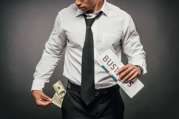 Cropped view of wealthy african american businessman holding business newspaper and looking at banknotes in pocket on dark background — Stock Photo