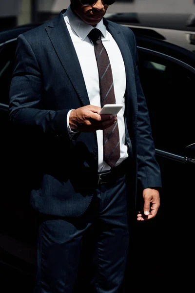 Vista recortada del hombre de negocios afroamericano en traje usando teléfono inteligente en el día soleado - foto de stock