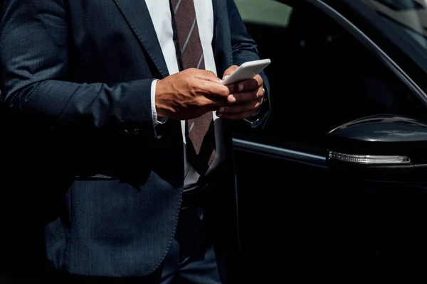 Vista ritagliata di uomo d'affari afro-americano in giacca e cravatta utilizzando smartphone nella giornata di sole vicino all'auto — Foto stock