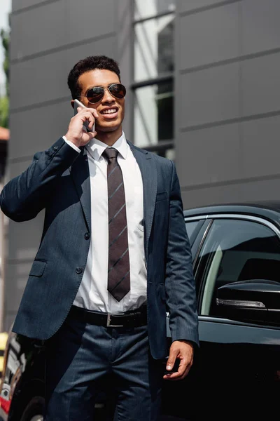 Hombre de negocios afroamericano en traje y gafas de sol hablando en el teléfono inteligente en el día soleado cerca del coche - foto de stock