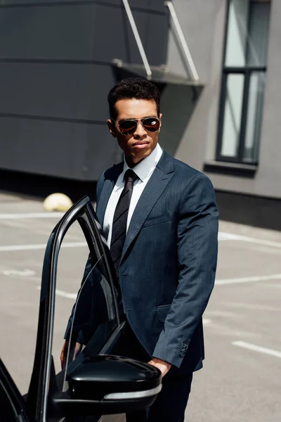 Homme d'affaires afro-américain en costume et lunettes de soleil marchant près de la voiture à la journée ensoleillée — Photo de stock