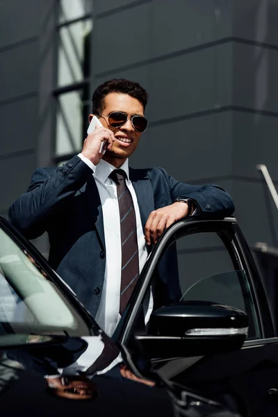 Homme d'affaires afro-américain en costume et lunettes de soleil parlant sur smartphone près de la voiture à la journée ensoleillée — Photo de stock