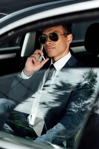 African american businessman in suit and sunglasses talking on smartphone in car at sunny day — Stock Photo