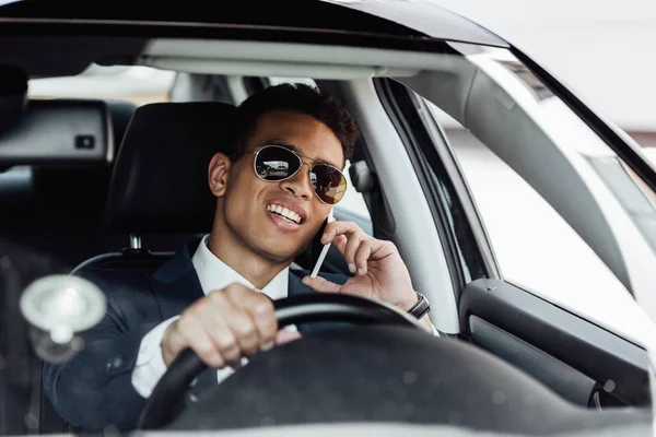 Un homme d'affaires afro-américain en costume et lunettes de soleil conduisant une voiture et parlant sur smartphone lors d'une journée ensoleillée — Photo de stock