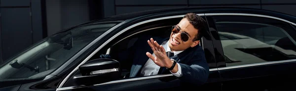 African american businessman in suit and sunglasses waving hand from car at sunny day, panoramic shot — Stock Photo