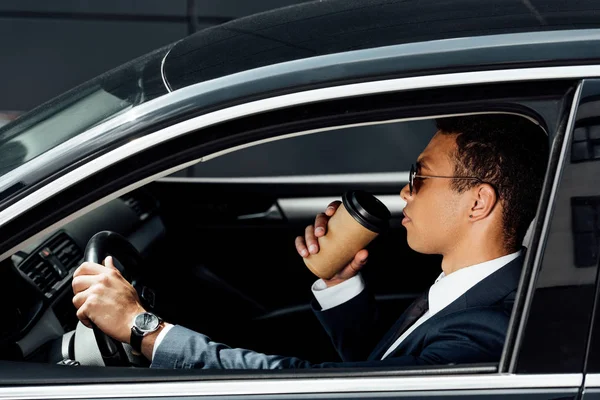 Vista lateral do empresário afro-americano de terno e óculos de sol dirigindo carro e bebendo café no dia ensolarado — Fotografia de Stock