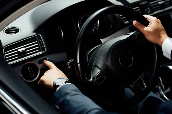 Cropped view of african american businessman in suit pushing button near steering wheel in car — Stock Photo